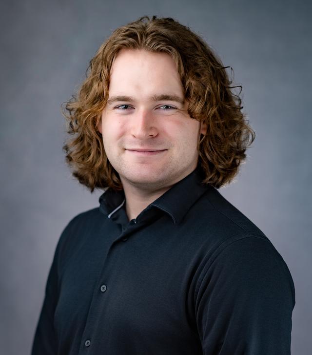 Brennan Keeney smiling in a dark button-up shirt, with shoulder-length curly red hair, in front of a gray studio backdrop