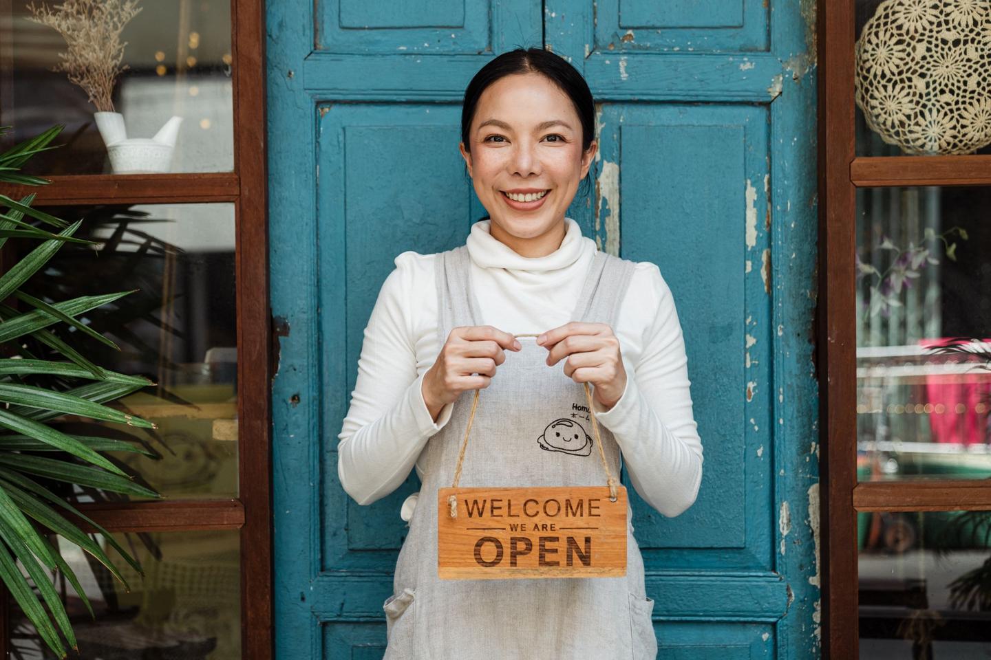 Smiling small business owner holding a We Are Open sign in front of her shop door