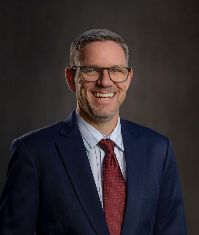 Lance Thomas smiling in a navy suit, light blue shirt, and red tie, wearing glasses, in front of a dark studio backdrop