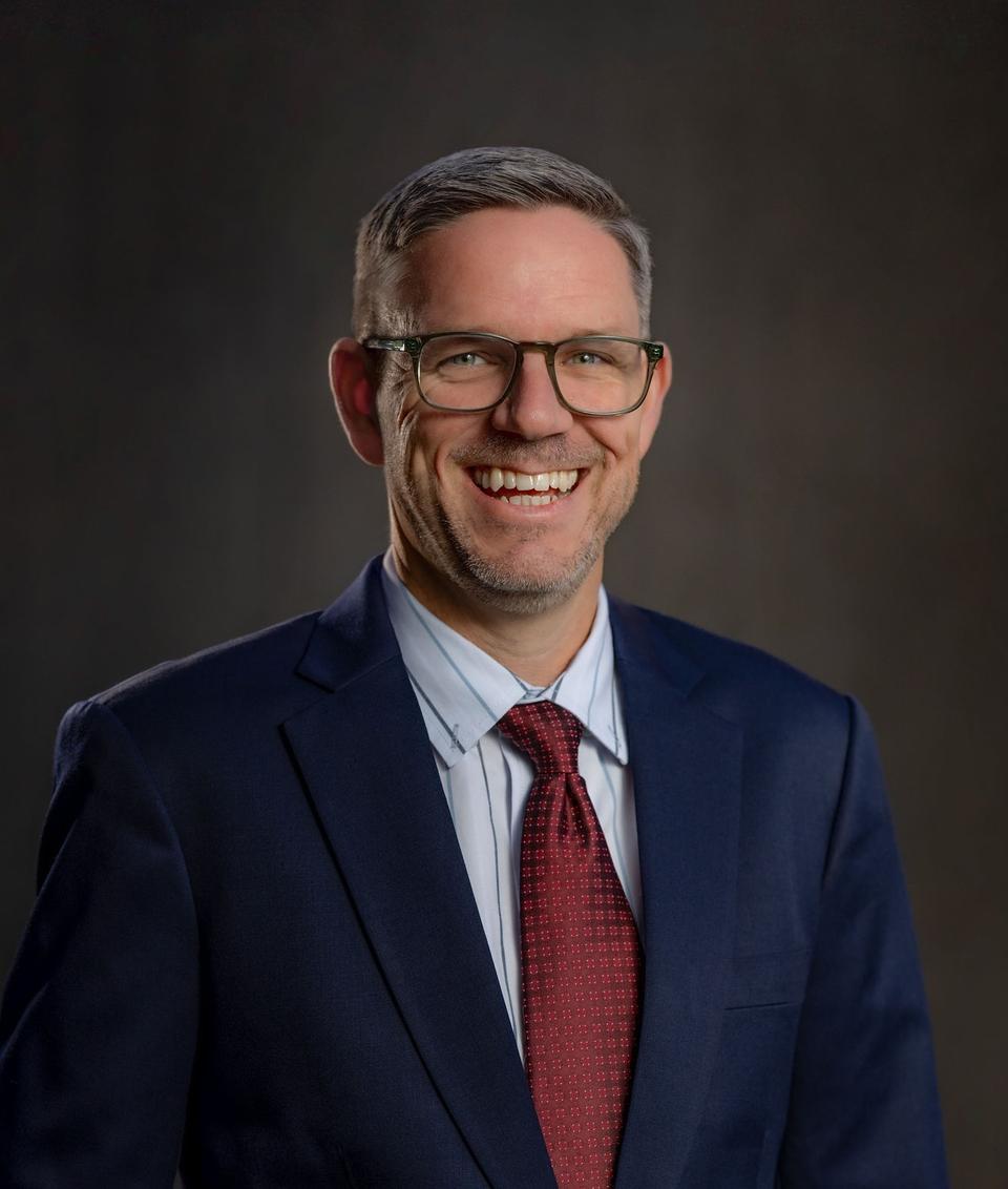 Lance Thomas smiling in a navy suit, light blue shirt, and red tie, wearing glasses, in front of a dark studio backdrop