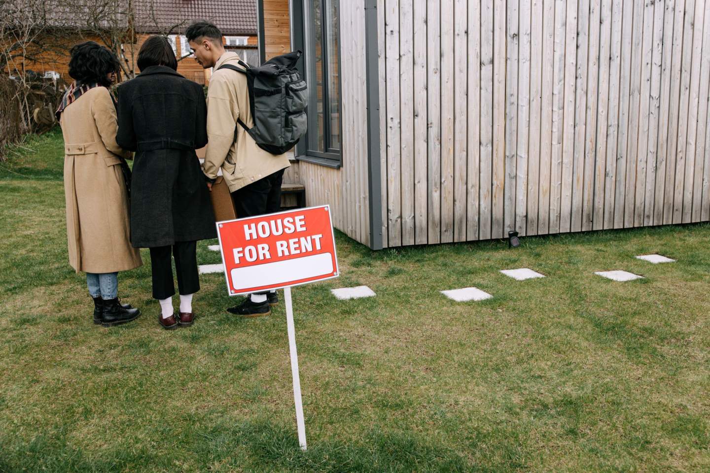 Three people standing on a lawn looking at a red House for Rent sign in front of a wood-sided house