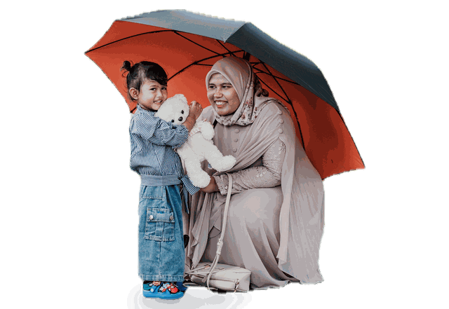 A smiling mother kneeling beside her young child and a white teddy bear under a large orange and gray umbrella